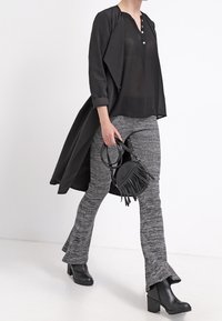Un long cardigan noir sur un top noir transparent, associé à un pantalon évasé gris texturé et des bottines noires. Un petit sac à franges complète le look.