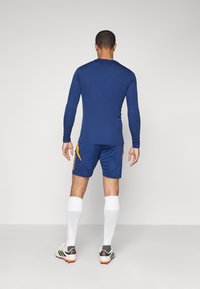 Meri panjang atletik navy dipadukan dengan celana pendek biru yang memiliki aksen kuning. Kaos kaki putih dan sepatu atletik hitam dengan detail merah.
