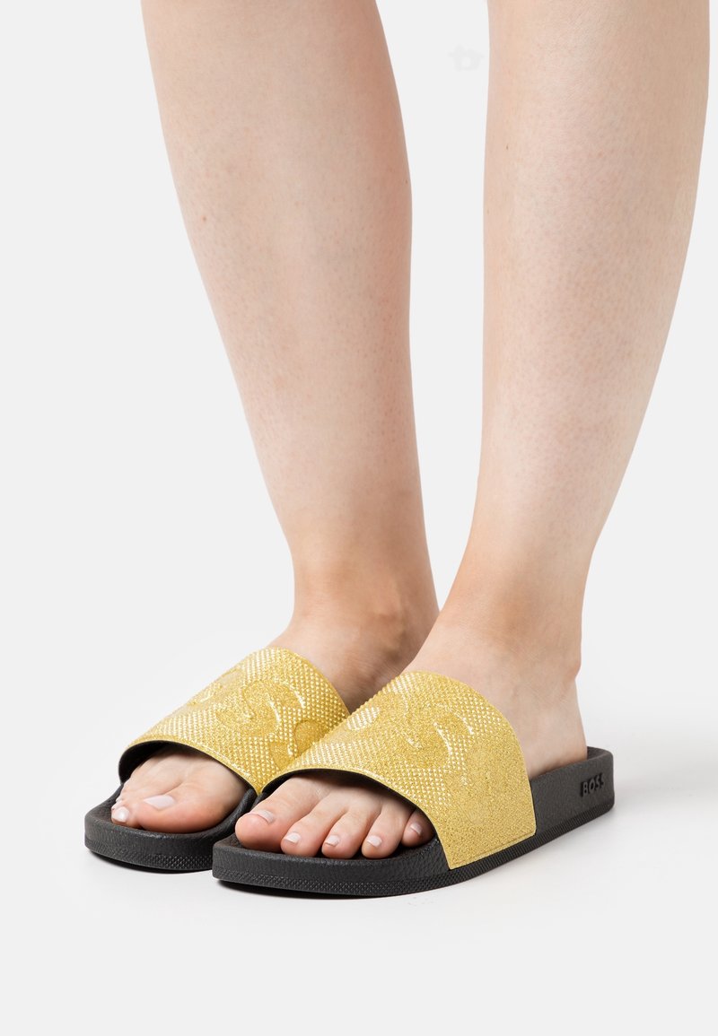 Pieds portant des sandales à semelle noire avec de larges brides dorées scintillantes embossées d'un logo sur fond blanc.