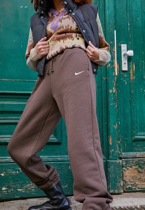 Persoon die bruine Nike sweatpants, zwarte laarzen, een gebreide trui met patroon en een zwarte puffer-vest draagt, staand voor een verweerde groene deur.