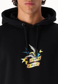 Svart huvtröja med en färgglad grafisk bild av en fågel med gula detaljer och ett banderoll som säger "SANTA CRUZ." Texturerad tyg med dragsnören.