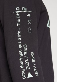 Chemise noire à manches longues avec texte graphique bleu clair et symboles sur la manche, présentant des motifs de style numérique dans un design moderne.