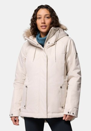 Donna che indossa un parka invernale beige con cappuccio foderato in pelliccia e sciarpa blu, in piedi contro uno sfondo chiaro e uniforme.