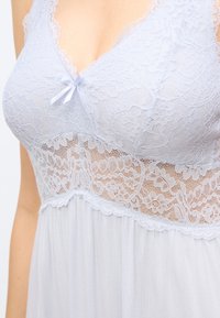 Λευκό bralette από δαντέλα με ανθικά μοτίβα, μικρό φιόγκο στο κέντρο και διάτρητη δαντέλα κάτω από το στήθος σε ανοιχτό ύφασμα.