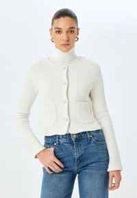 Valkoinen neulecardigan, jossa on pyöreä kaula-aukko, kaksi taskua ja kultaiset napit. Käytetään poolopaidan päällä, yhdistettynä sinisiin farkkuihin.
