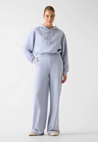 Sweat à capuche bleu clair avec un logo texturé, assorti à un pantalon à jambes larges. Les deux articles sont confectionnés dans un tissu doux et offrent une coupe décontractée.