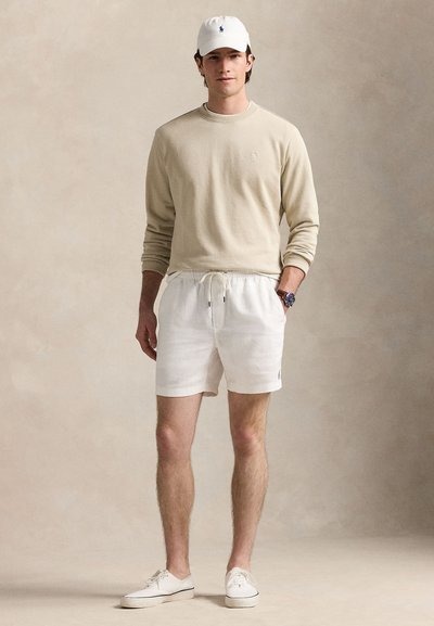 Camisa bege de manga comprida, calções brancos com cordão, boné branco e sapatos brancos. Os calções têm bolsos laterais e um pequeno detalhe de logótipo.