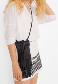 Mujer vestida con una blusa blanca abotonada y una falda blanca con estampado, llevando un bolso cruzado de cuero negro con flecos largos.