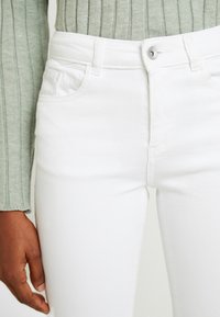 En person som bär vita jeans och en ljusgrön ribbad tröja, med vänsterhanden avslappnad vid sin sida.