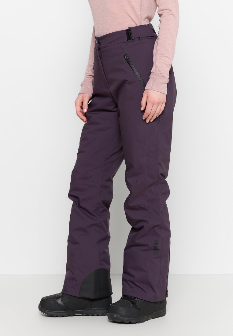 Person in lila Skihose mit seitlichem Reißverschluss und schwarzen Schneestiefeln, stehend vor einem weißen Hintergrund.