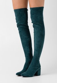 Türkisfarbene Wildleder-Stiefel über dem Knie mit Blockabsätzen und seitlichem Reißverschluss, getragen auf nackten Beinen vor einem schlichten weißen Hintergrund.