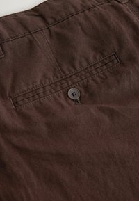Gros plan d'un pantalon en tissu marron montrant une poche arrière avec un seul bouton foncé et des coutures détaillées.