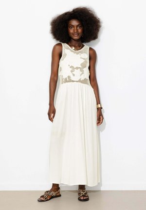 Femme aux grands cheveux bouclés portant une robe maxi blanche sans manches avec un corsage en résille fleurie et des sandales à imprimé léopard, debout contre un mur blanc.