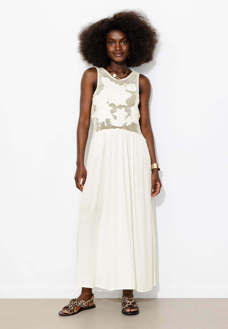 Femme aux grands cheveux bouclés portant une robe maxi blanche sans manches avec un corsage en résille fleurie et des sandales à imprimé léopard, debout contre un mur blanc.
