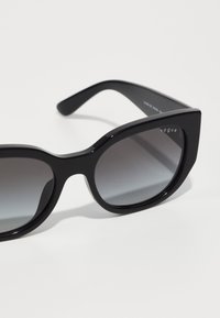 Black sunglasses with a geometric design, glossy frame, gradient lenses, and "vogue" branding etched on the side.