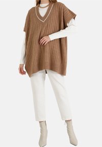 Beżowa, dzianinowa, oversize kamizelka o prążkowanej fakturze i dekolcie w serek, zestawiona z białą koszulą z długim rękawem oraz kremowymi, dopasowanymi spodniami.