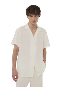 Camicia a maniche corte con colletto in tessuto bianco leggero e texture, con chiusura a bottoni e vestibilità rilassata. Presenta un design casual e aperto.
