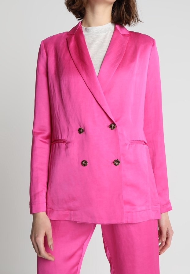 Femme portant un blazer en satin rose vif à double boutonnage avec un pantalon assorti et un t-shirt blanc à col rond, debout devant un fond uni.