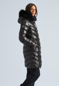 Melna pūkā vecmāte ar spīdīgu apdari, ar kažokādas apdari kapuci, ar rāvējslēdzēja kabatām un diagonālām šuvēm, kas piešķir tekstūru.