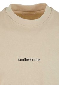 Kortærmet beige t-shirt i bomuld med rund hals og sort "AnotherCotton" logo på brystet. Glat tekstur, minimalistisk design.