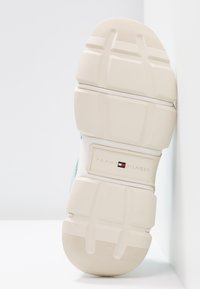 Semelle de basket couleur crème avec des rainures profondes et le logo "Tommy Hilfiger" au centre sur fond blanc.