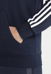 Námořnická modrá mikina s klokáním kapsou, žebrovanými manžetami a bílými pruhy na rukávech pro kontrast.