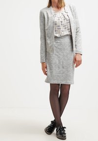 Blazer gris texturizado sobre una blusa con estampado, combinado con una falda lápiz gris a juego. Zapatos de charol negros completan el atuendo.