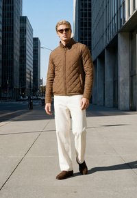 Un uomo che indossa occhiali da sole, una giacca trapuntata marrone, pantaloni bianchi e scarpe marroni cammina su un marciapiede della città tra edifici alti.