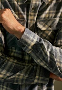 Camisa de franela en cuadros azules y beige. Presenta un puño abotonado con herrajes negros en contraste y un tejido texturizado.