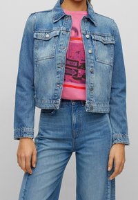 Žena v světle modré džínsové bundě a džínách, pod kterými má růžové tričko s grafikou, stojící s rukama u těla.