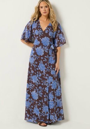 Kobieta ubrana w brązową sukienkę kopertową w niebieski kwiatowy wzór, z krótkimi szerokimi rękawami i paskiem w talii, stojąca na tle jasnej, gładkiej ściany.