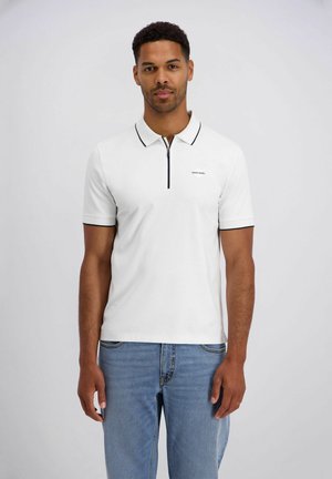 Ein Mann steht frontal und trägt ein weißes, kurzärmeliges Poloshirt mit schwarzen Akzenten und blaue Jeans vor einem einfarbigen grauen Hintergrund.