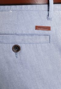 Niebieskie dżinsy z subtelnym wzorem jodełki. Posiadają brązową skórzaną metkę oraz okrągły ciemny guzik na przedniej kieszeni.