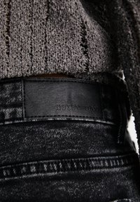 Zwarte denim jeans met een gemêleerde, grijze gebreide trui die gedeeltelijk zichtbaar is. Zwarte leren label met de tekst "NOISY MAY" op de taille.