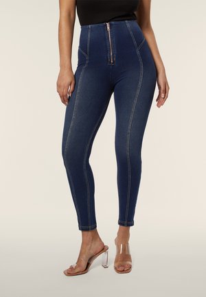 Jeans skinny in denim a vita alta di colore blu scuro con cerniera frontale e cuciture gialle a contrasto. Abbinati a sandali con tacco trasparenti.