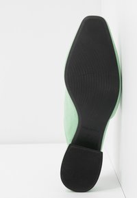 Semelle d'une chaussure vert menthe avec une outsole en caoutchouc noir et un petit talon, reposant verticalement contre un angle de mur et de sol blancs.