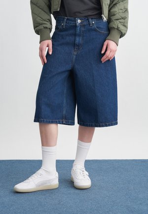 Isik, kes kannab põlveni ulatuvaid laiu teksapükse, valgeid sokke, valgeid tenniseid, musta särki ja rohelist tikandiga jopet, seisab sinisel vaibal.