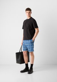 Svart t-skjorte med kort erme og lomme, blå rutete shorts, svarte loafers og en svart tote bag. Glatt stoff med en avslappet passform.