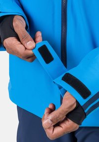 Chaqueta impermeable azul con puños de Velcro negros y una cremallera. Presenta una textura suave y un diseño ajustado, mostrando manos ajustando el puño.