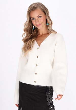Cardigan bianco a coste con scollo a V, dotato di bottoni dorati lungo il davanti e maniche lunghe. Abbinato a una gonna nera con dettagli decorativi.