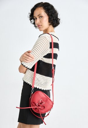 Red leather crossbody bag with lace-up detail, rounded shape, adjustable strap, and smooth texture, paired with a striped knit top and black skirt.