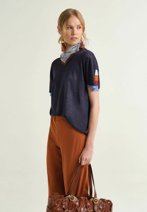 Donna che indossa una maglietta a maniche corte blu navy, pantaloni color ruggine, sciarpa a fantasia, orecchini a nappina, che tiene in mano una borsa di pelle marrone con ritagli floreali.