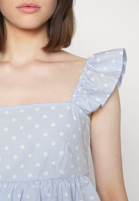 Woman wearing a light blue dress with white polka dots and ruffled shoulder straps, showing neck, collarbone, and upper arm.