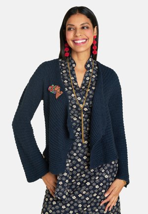 Navy knitted cardigan with draped open front, textured fabric, and a floral brooch. Paired with a patterned dress featuring white flowers.