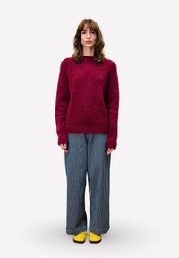 Eine Frau steht und trägt einen dunkelroten Pullover, weite blau-weiße gestreifte Hosen und leuchtend gelbe Schuhe vor einem einfachen hellen Hintergrund.