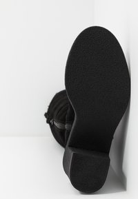 Vue de la semelle d'une botte en daim noir avec un talon bloc, une semelle antidérapante texturée et une fermeture éclair latérale, positionnée contre un coin blanc.