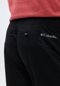 Κοντινό πλάνο ενός ατόμου που φοράει μαύρο παντελόνι Columbia με φερμουάρ στην πίσω τσέπη και κόκκινο πουκάμισο που έχει μπει μέσα στο παντελόνι.