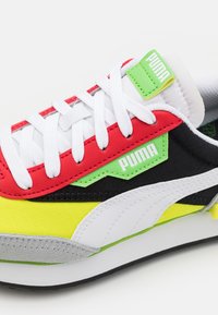 Mehrfarbiger Sneaker mit einem rot-schwarzen Obermaterial, gelben Akzenten, weißen Schnürsenkeln und einem grünen Logoreiter. Überall strukturierte Materialien.