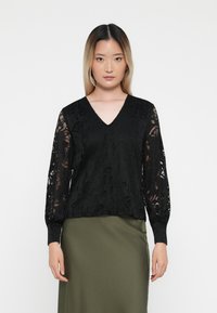 Μαύρη μπλουζοκόντη με λουλούδια από δαντέλα, με V-λαιμό, μακριά διαφανή μανίκια και υφή στο σώμα; συνδυασμένη με μια ελιά πρασινιστή φούστα.