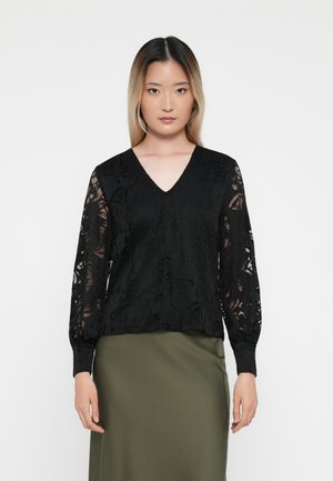 Blusa in pizzo floreale nero con scollatura a V, maniche lunghe trasparenti e corpo testurizzato; abbinata a una gonna verde oliva.